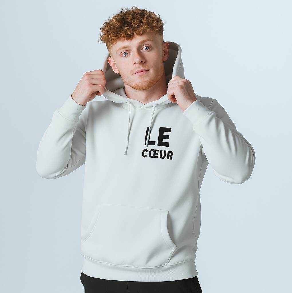 Young man with curly red hair and fair skin wearing a white hoodie printed with "LE CŒUR," pulling the hood up and looking at the camera against a pale blue background.