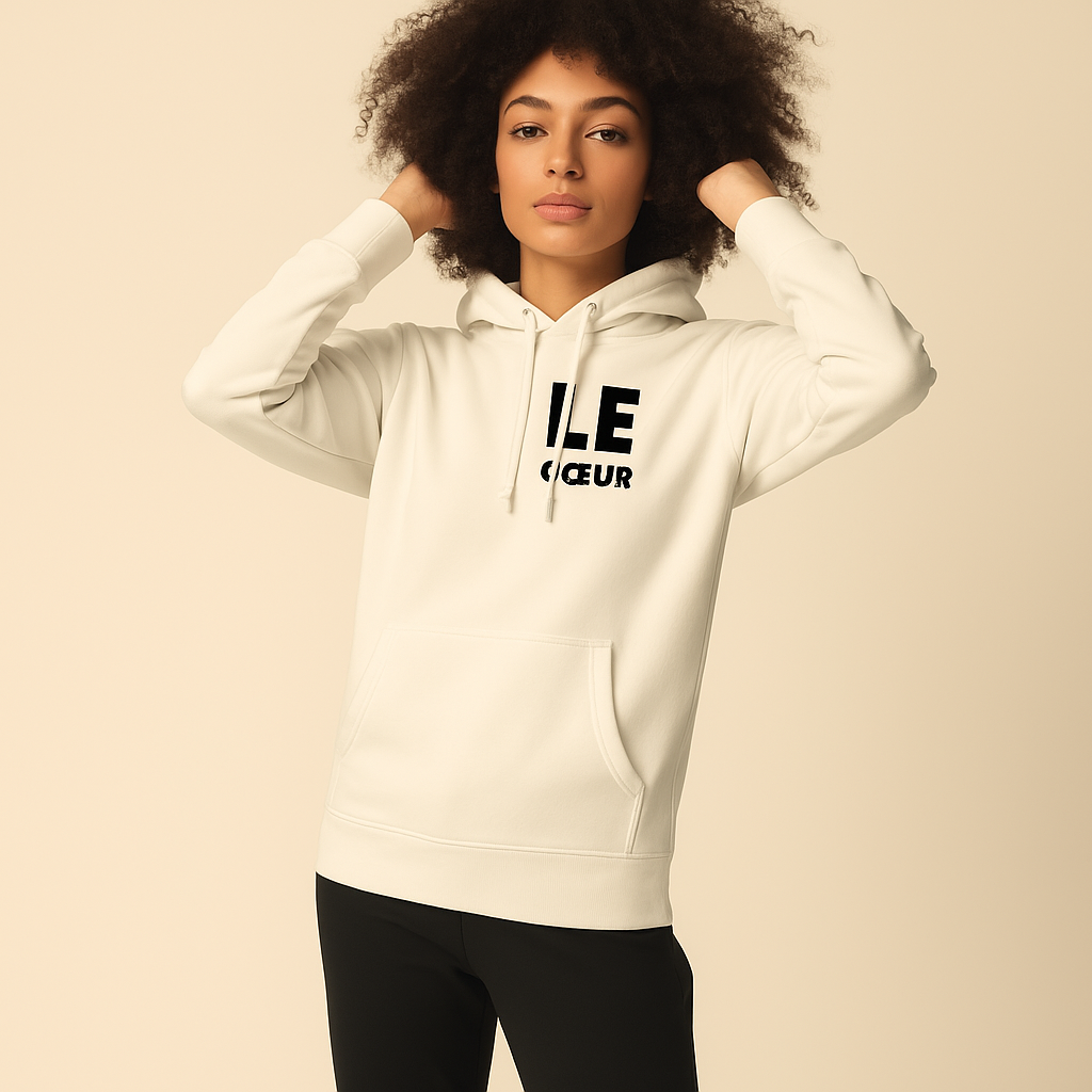 Woman with a voluminous afro standing against a plain beige background, wearing a cream hoodie with bold black 'LE' lettering and black pants, hands raised to her hair and a calm expression.