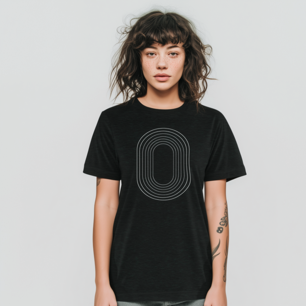 Young woman with wavy dark hair, bangs and a freckled face standing against a light gray background, wearing a black T-shirt printed with concentric white oval lines and showing forearm tattoos.