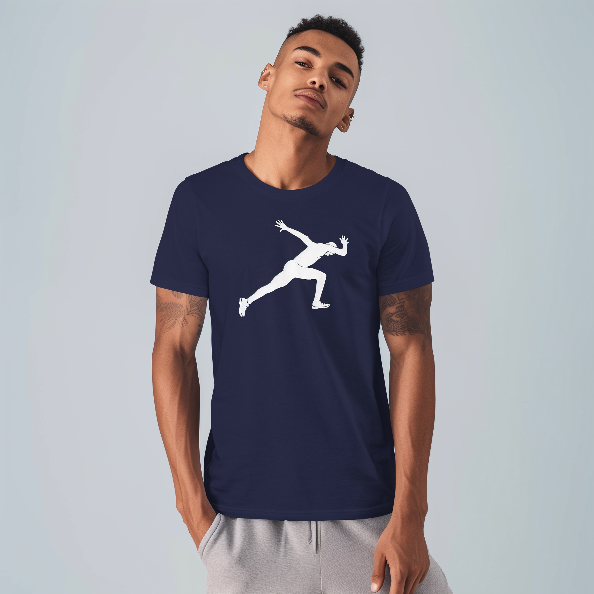 Man standing against a pale gray background wearing a navy T-shirt printed with a large white silhouette of a running figure, hands in his pockets, head tilted and forearm tattoos visible.